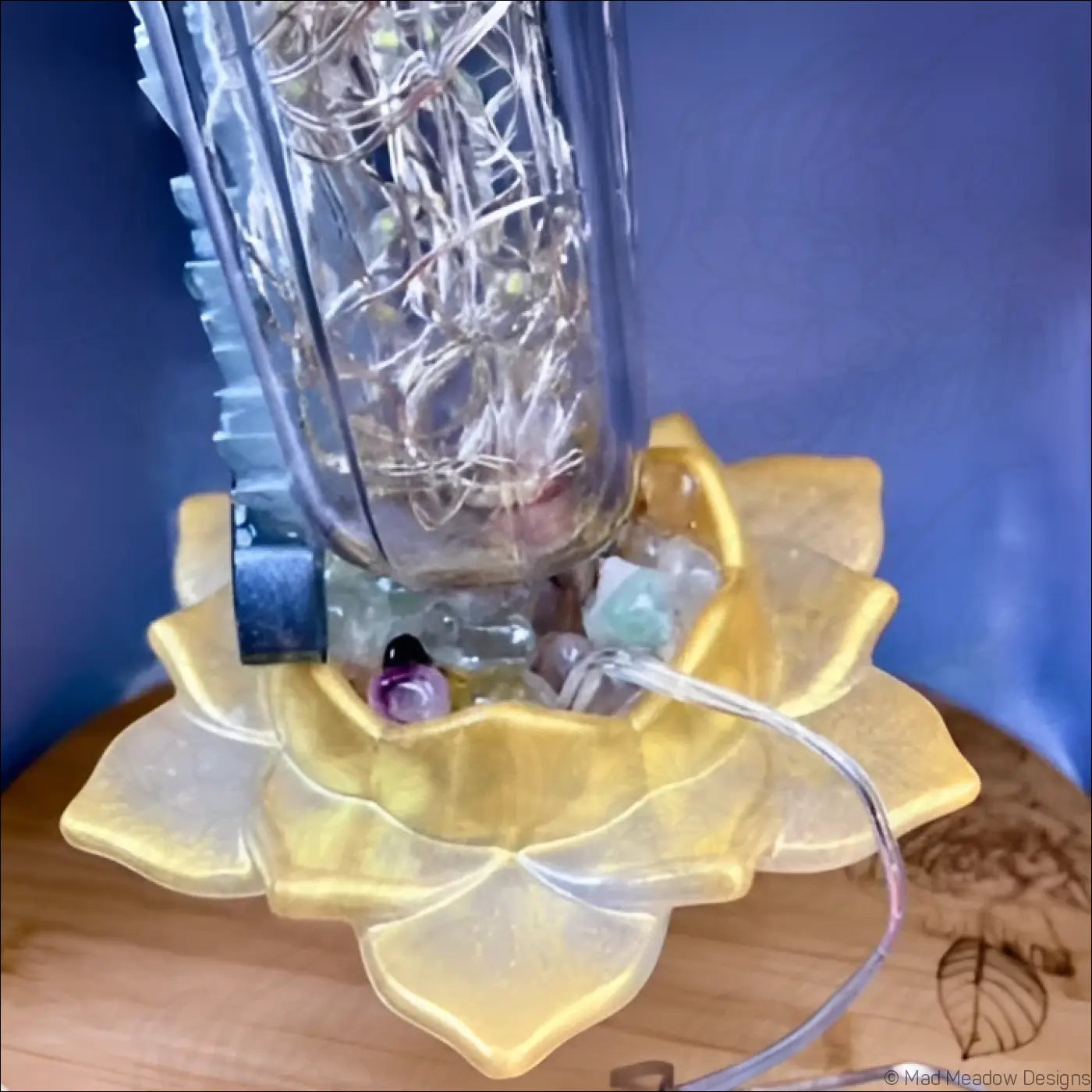 A clear glass apothecary bottle lantern with intricate wirework on a yellow lotus base with colorful gemstones