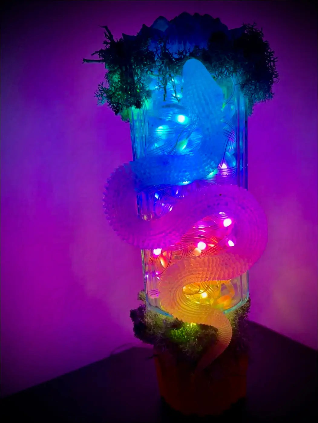 A glowing multi-colored plastic snake sculpture with lights coiled around a transparent antique glass apothecary bottle usb light with moss and crystals