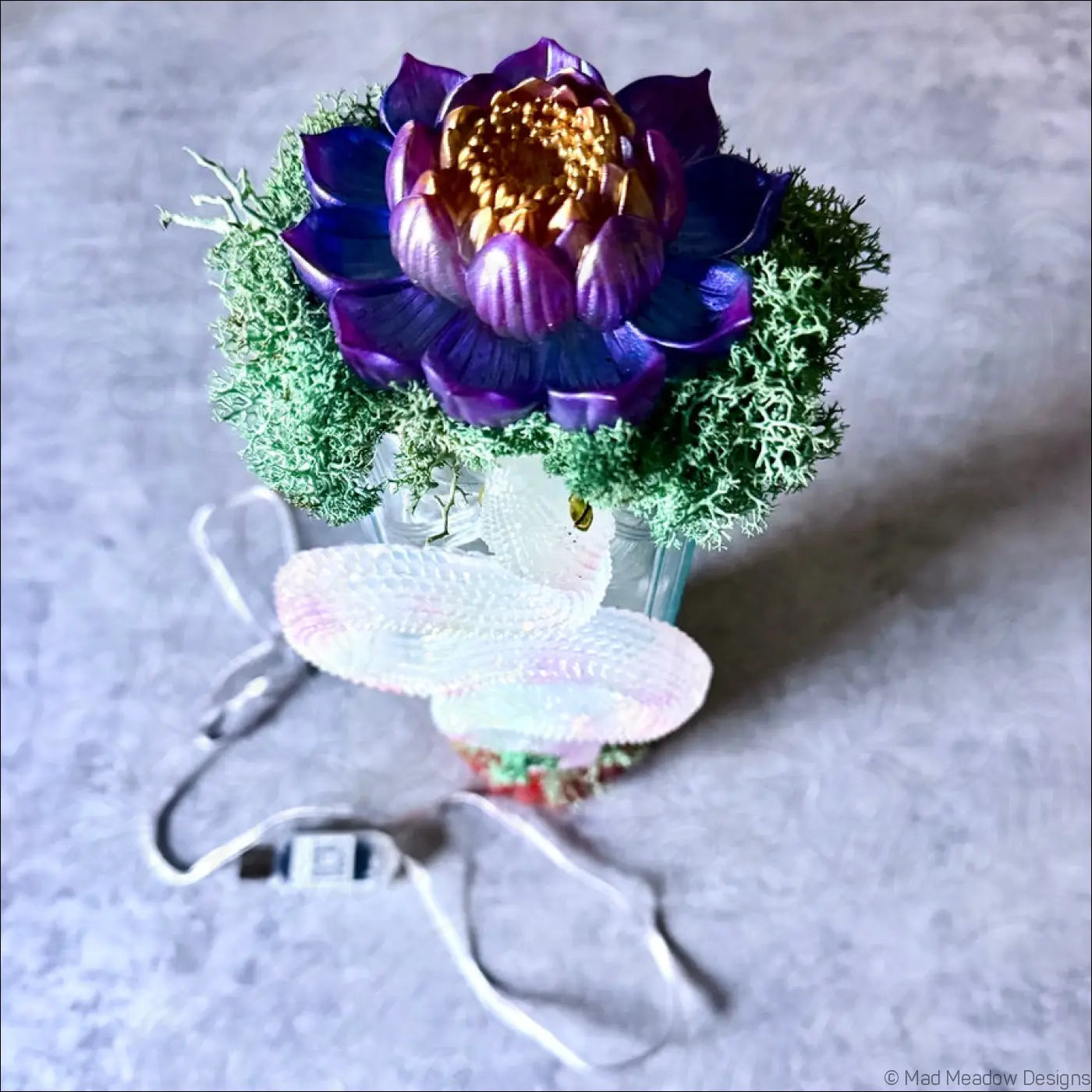 A decorative purple and gold lotus flower centerpiece with green moss in a glass vase on an antique glass apothecary bottle usb light