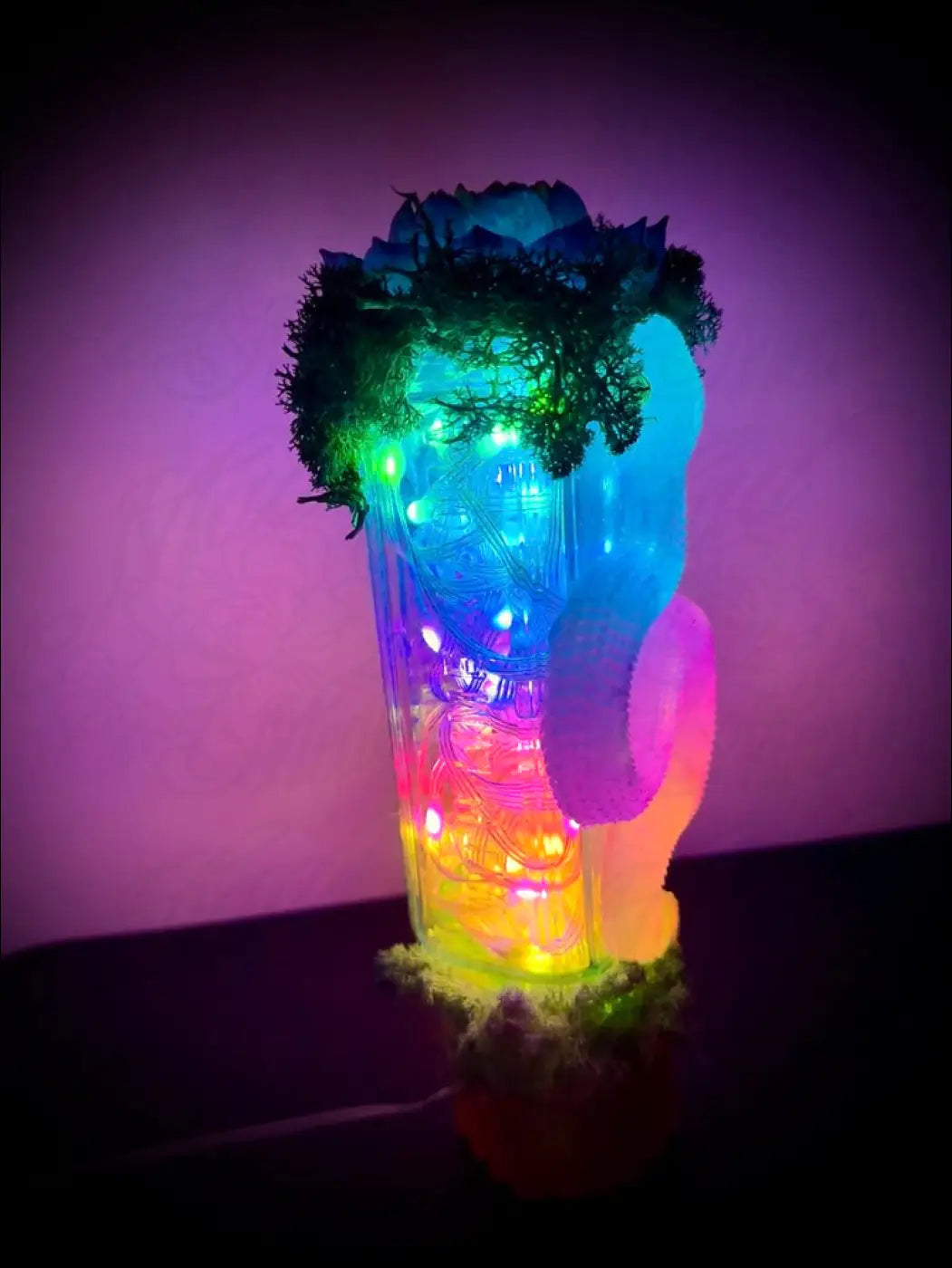 A translucent rainbow-colored led lamp shaped like a beer mug with glowing lights and greenery on an antique glass bottle