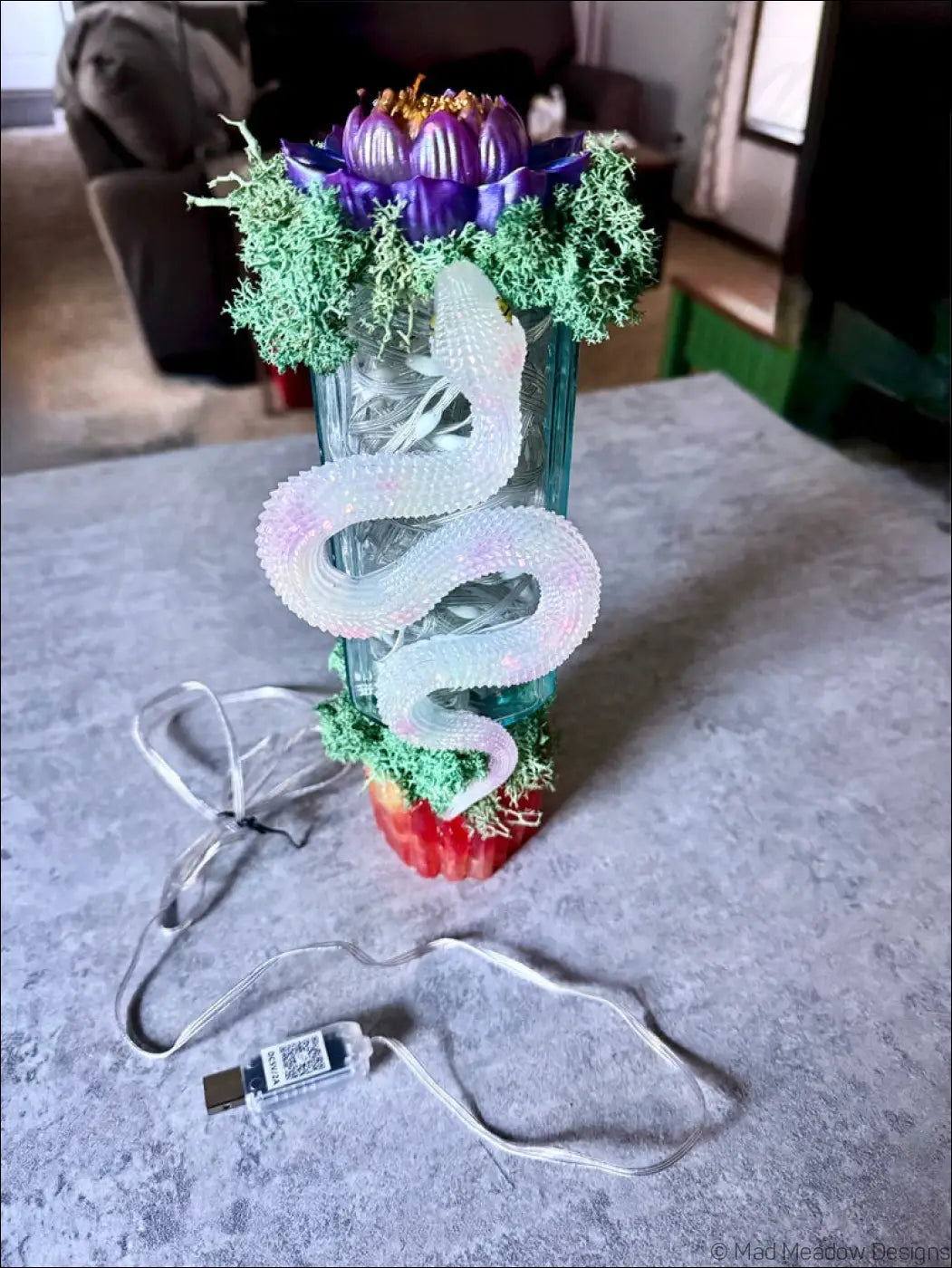 A glowing white snake sculpture coiled on a glass vase with purple lotus flowers and green moss usb light