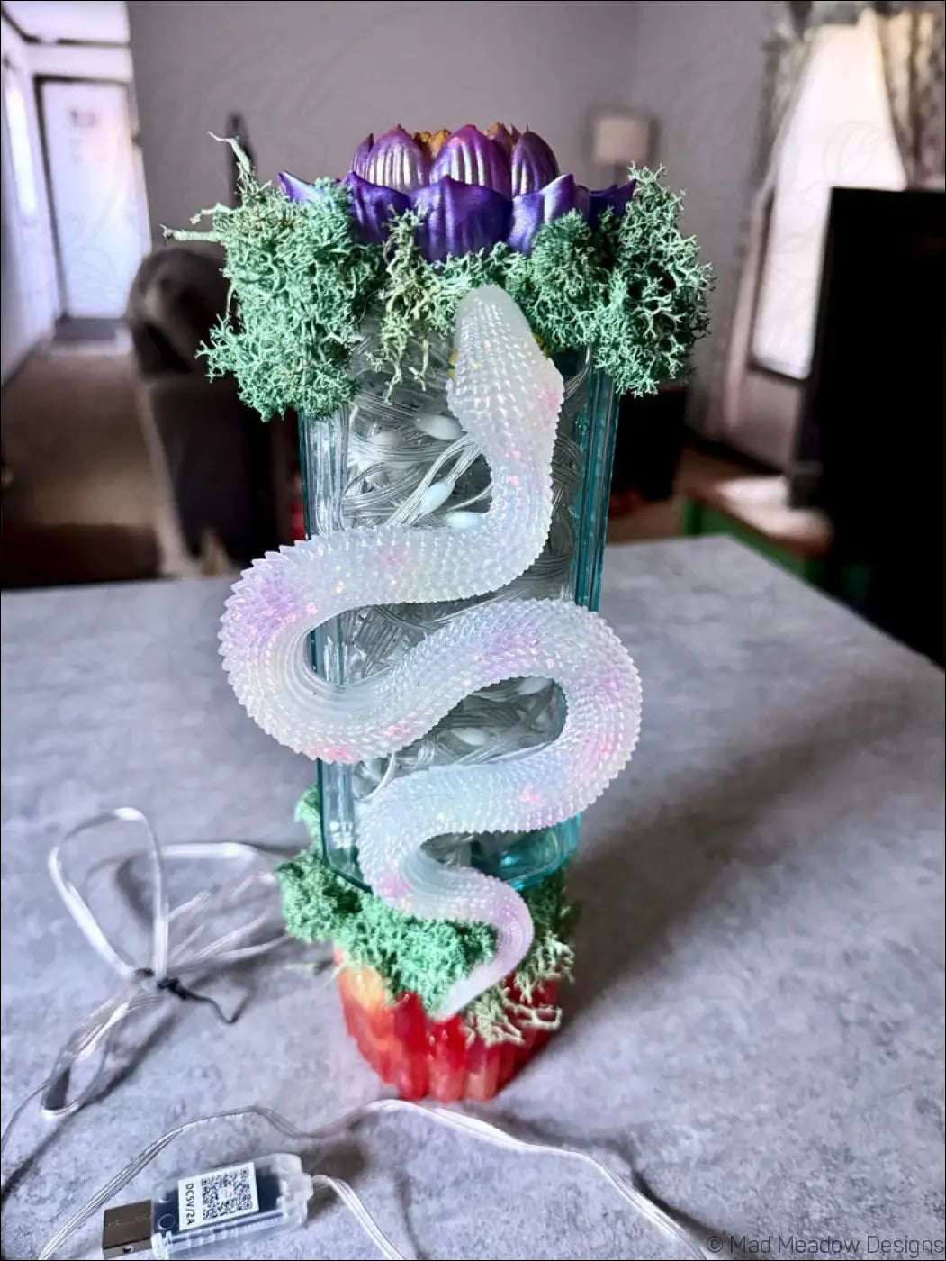 A translucent iridescent snake sculpture coiled on a glass apothecary vase with green moss and a purple lotus flower usb light