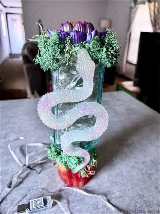 A translucent iridescent snake sculpture coiled on a glass apothecary vase with green moss and a purple lotus flower usb light