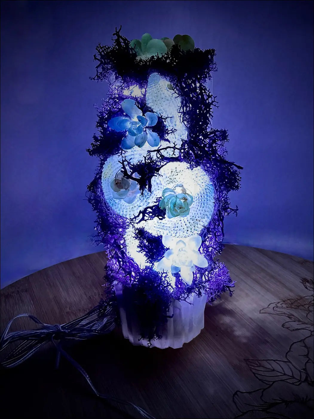 A glowing skull-shaped usb lamp with blue and white succulents nestled in pastel purple moss and crystals
