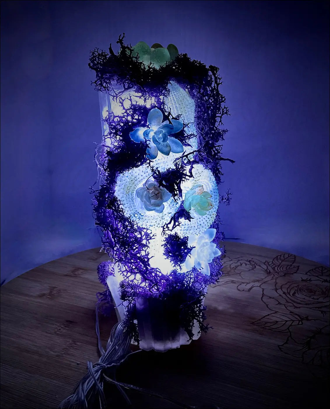 A glowing skull lamp on an antique glass bottle with green stones, blue and white succulents, and pastel purple moss