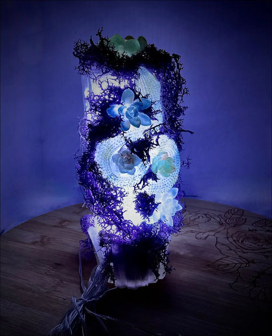 A glowing skull lamp on an antique glass bottle with green stones, blue and white succulents, and pastel purple moss