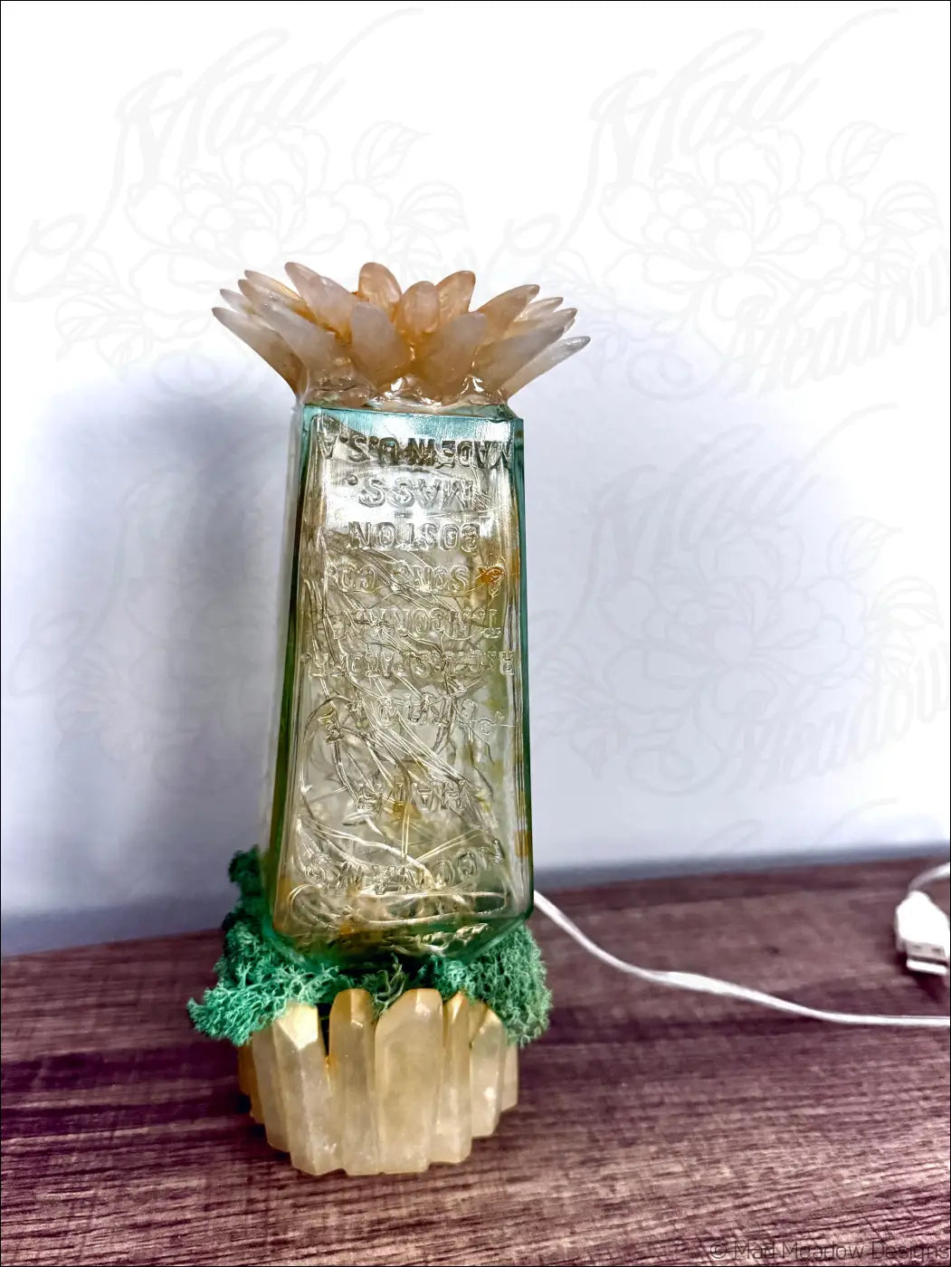 A translucent green glass hair petrole bottle with embossed text and frosted petal top on moss and crystal base