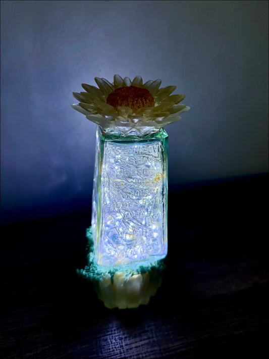 A translucent green glass hair petrole bottle lantern with etched patterns topped by a yellow and orange sunflower