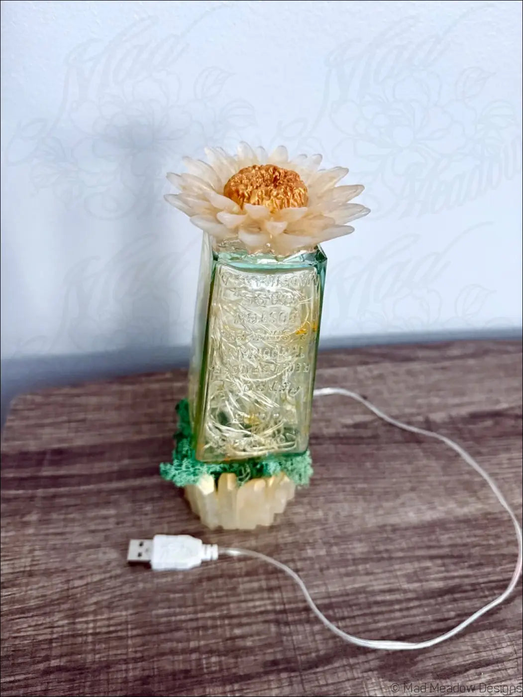 A translucent green glass hair petrole bottle with etched patterns topped by a dried cream daisy on green base