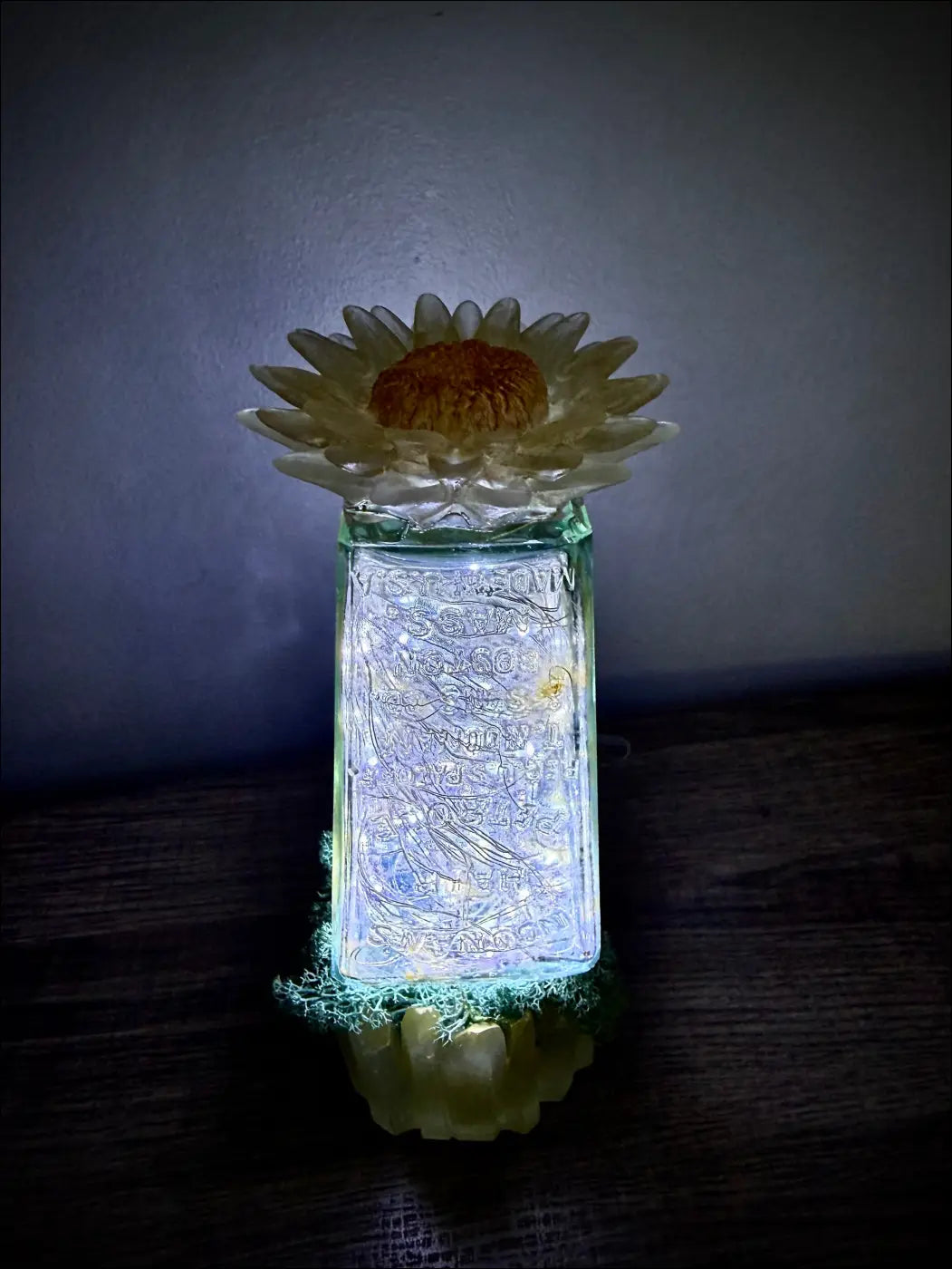 A translucent hair petrole bottle glass lantern with etched designs lit by soft white lights and topped with a cream daisy