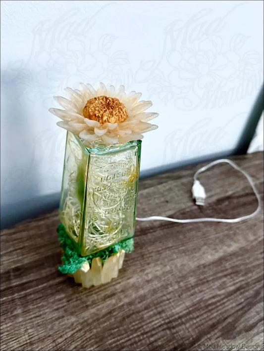 A cream dried flower with a golden center on a vintage green glass hair petrole bottle with etched patterns and crochet base